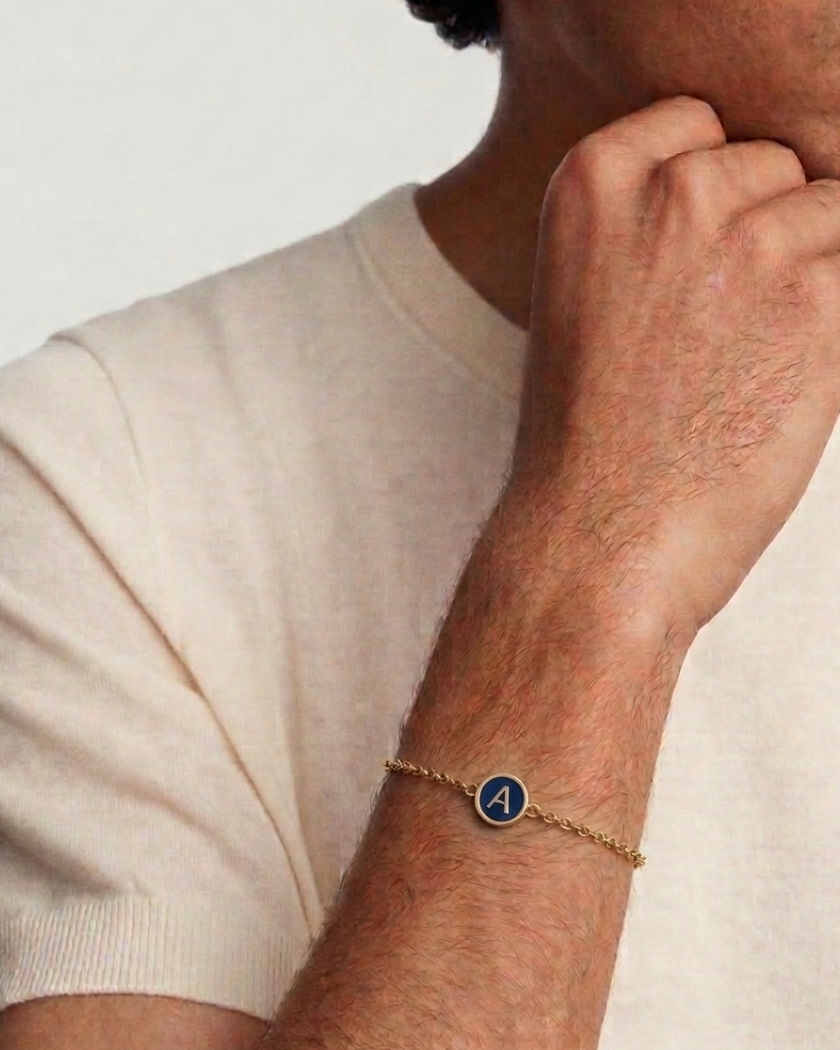 Man wearing a gold bracelet with a blue charm on a plain background