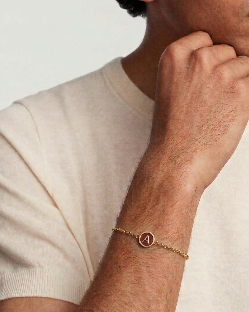 Man wearing a gold bracelet with a letter 'A' charm on a plain background