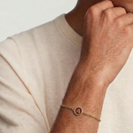 Man wearing a gold bracelet with a letter 'A' charm on a plain background