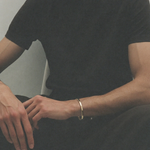 A man wearing a simple gold bangle bracelet on his wrist, dressed in a black t-shirt and black trousers while sitting.